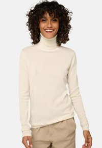 Frau mit lockigem Haar, die lächelt, trägt einen cremefarbenen Rollkragenpullover und beige Hosen und steht vor einem schlichten hellen Hintergrund.