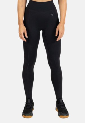 Persona con leggings negros de talle alto y zapatillas deportivas negras con suelas marrones, de pie frente a un fondo liso.