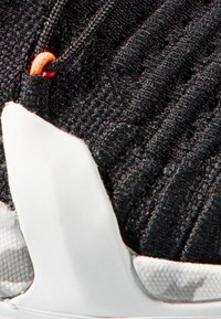 Svart idrettssko med en teksturert overdel med tynne svarte stoffer, hvit såle og en oransje detalj nær snøringene.