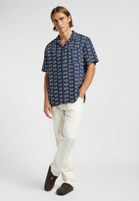 Chemise à manches courtes marine avec un motif géométrique blanc, design à boutons, et poche, accompagnée de pantalons couleur crème et de sabots marron.