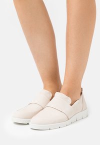Zapatos deslizantes de color beige claro, hechos de suave gamuza, que cuentan con una ancha banda elástica en la parte delantera y una suela de goma blanca con un diseño texturizado.