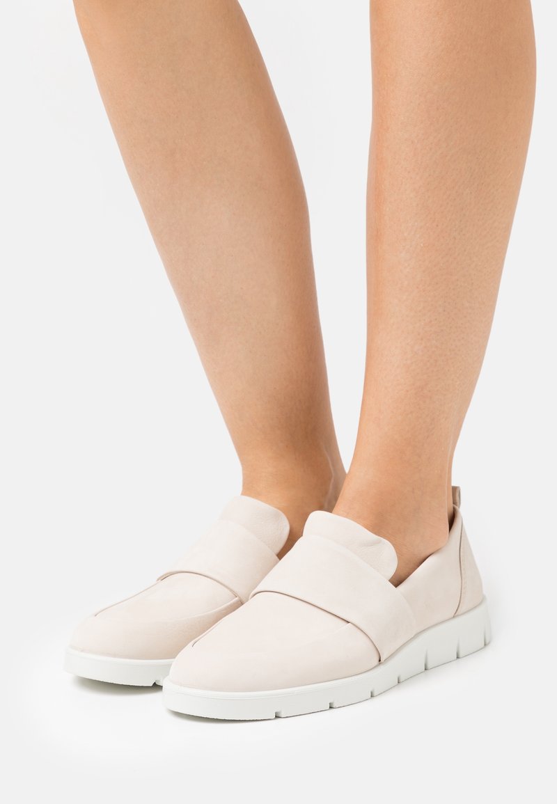 Zapatos deslizantes de color beige claro, hechos de suave gamuza, que cuentan con una ancha banda elástica en la parte delantera y una suela de goma blanca con un diseño texturizado.