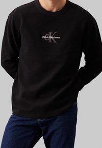 Černý sweatshirt s dlouhým rukávem a strukturovanou tkaninou. Obsahuje malé vyšívané logo v bílé a šedé barvě. Vybaveno modrými džíny.