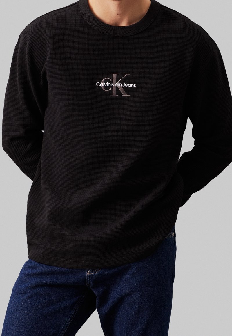 Černý sweatshirt s dlouhým rukávem a strukturovanou tkaninou. Obsahuje malé vyšívané logo v bílé a šedé barvě. Vybaveno modrými džíny.