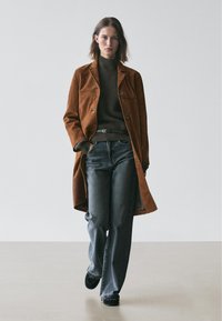 Bruine suède lange jas over een donker geribbelde coltrui, gecombineerd met wijde blauwe jeans. De outfit heeft minimale accessoires en monochrome tinten.