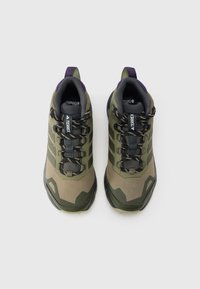 Zöld és szürke túracipők texturált felsőrésszel, gumitalppal, kiemelkedő fűzőrendszerrel és lila részletekkel a saroknál.