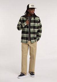 Green and black plaid flannel shirt, gray T-shirt, beige pants, and navy shoes. Hat has a beige front panel and logo.