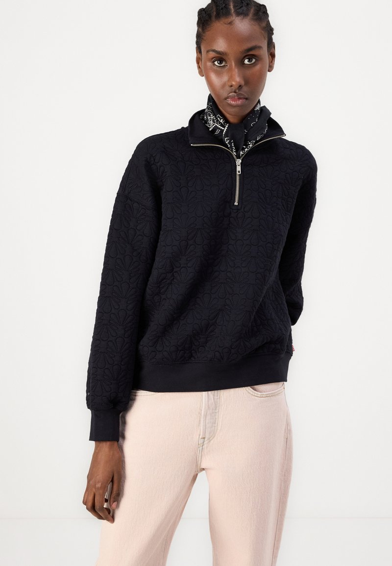 Sudadera negra con cremallera y textura floral, cuello alto y puños acanalados, combinada con pantalones rosa claro. Presenta un detalle de cremallera decorativa.