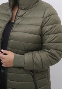 Oliivinvihreä toppatakkinen untuvatakki, jossa on korkea kaulus, vetoketjullinen etuosa ja resorireunukset hihoissa. Takissa on vaakasuuntainen tikattu kuviointi ja teksturoitu pinta.