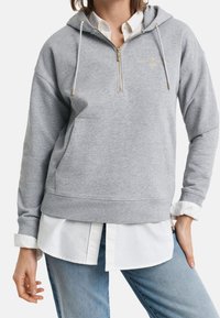 Grijze hoodie met ritssluiting en een goudkleurig embleem, voorzien van een capuchon met trekkoord, een zak aan de voorkant en een geribbelde zoom, gedragen boven een wit shirt.