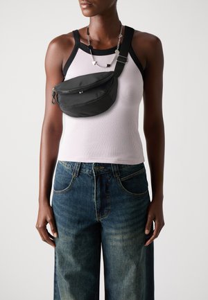 Geantă neagră din nailon tip crossbody, cu închidere cu fermoar, purtată peste un top alb din țesătură cu striuri, asortată cu blugi bleu largi. Accesorii argintii prezente.