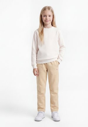Ung pige stående, iført en lys beige sweatshirt, brune joggingbukser med et lille logo og hvide sneakers, mod en ensfarvet hvid baggrund.