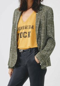 Grönmönstrad blazer över en gul V-ringad t-shirt med "STOCK" tryckt i svart. Svarta byxor med ett silverbälte.