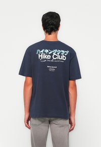 Hombre que lleva una camiseta azul marino con el texto "Hike Club Sakura" en japonés, de pie frente a una pared blanca, vistiendo pantalones gris claro.