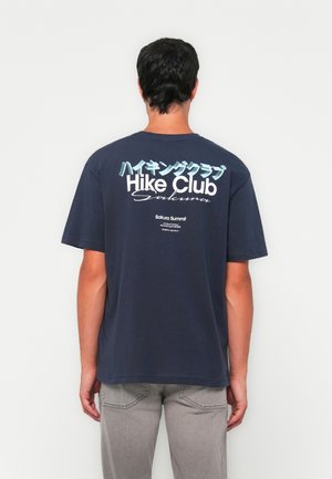 Moški nosi temno modro majico "Hike Club Sakura" z japonskim besedilom, stoji proti belemu zidu in nosi svetlo siva hlače.