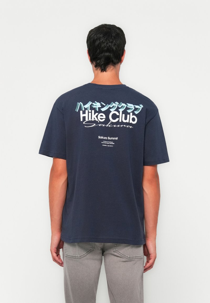 Hombre que lleva una camiseta azul marino con el texto "Hike Club Sakura" en japonés, de pie frente a una pared blanca, vistiendo pantalones gris claro.