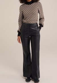 Brun och svart mönstrad tröja med hög krage, parat med svarta byxor i faux läder med breda ben. Strukturerad stickning och precisa sömnadsdetaljer.