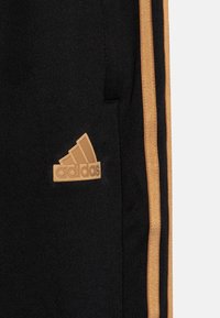 Melnas sporta bikses ar faktūru, ar zeltītām sānu svītrām un reljefu bežā krāsā Adidas logo kreisajā kabatas pusē.
