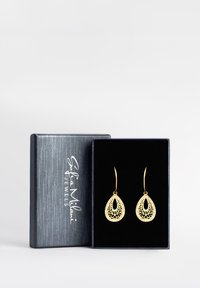 Guldiga tårformade örhängen med intrikata utskärningar, visade i en svart sammetsfodrad låda, med ett silverstämplat varumärke på locket.