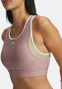Woman wearing a mauve and beige sports bra with white trim, featuring the Under Armour logo on the chest, cropped at the neck and waist.