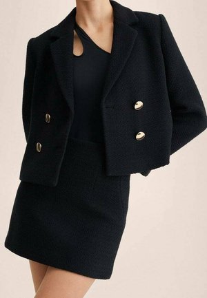 Kvinde iført en sort tekstureret cropped blazer med guldknapper og matchende højtaljet mininederdel over en sort top med udskæring.