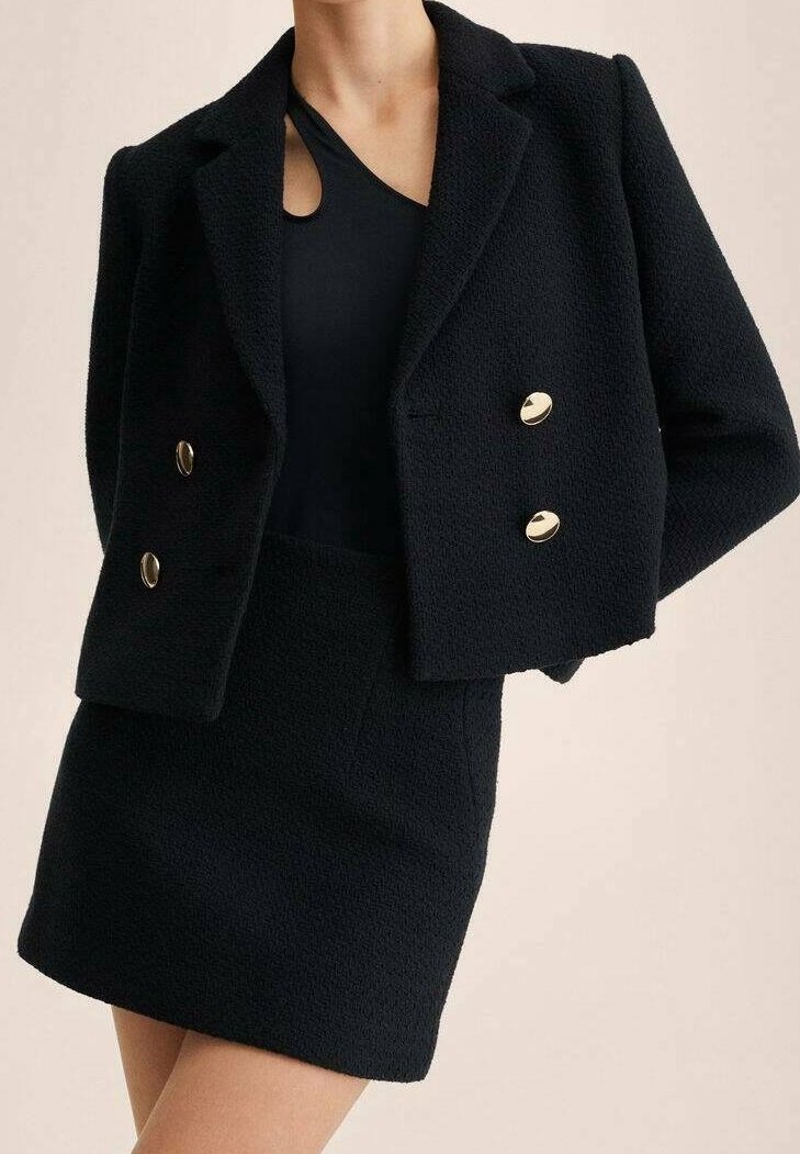 Femme portant un blazer court texturé noir avec des boutons dorés et une mini-jupe taille haute assortie sur un top noir avec une découpe.