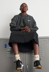 Zwarte denimrok en lichtgrijze jas op een zittend persoon. Zwarte sneakers met witte en tan accenten. Witte sokken zichtbaar. Grijze kluizen op de achtergrond.