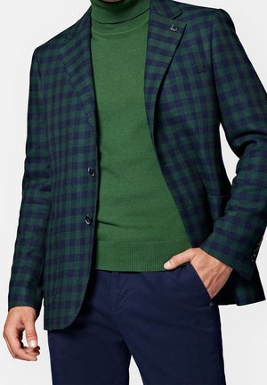 Man met een groene coltrui, een marineblauw en groen geruit colbert en een marineblauwe broek met één hand in de zak.