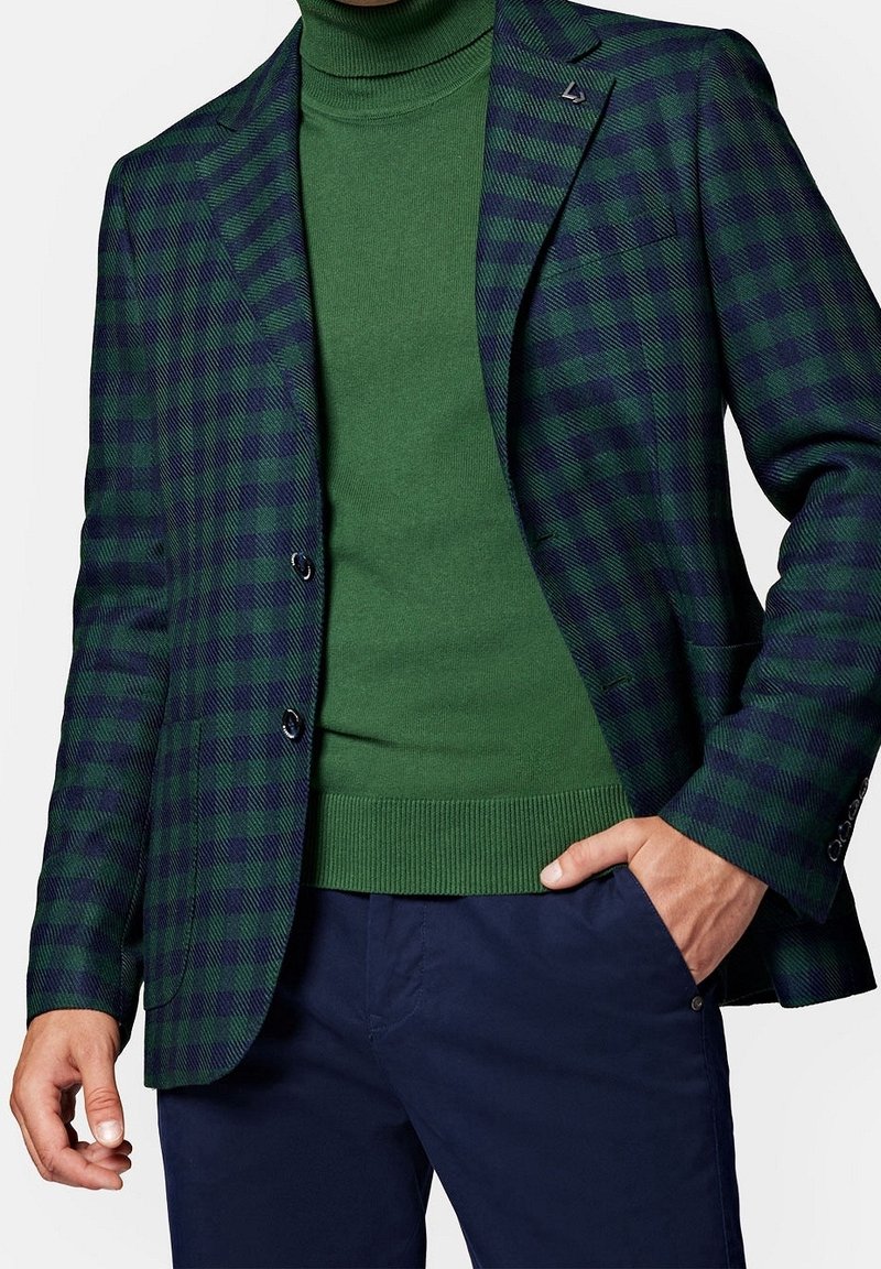 Homme portant un col roulé vert, un blazer à carreaux bleu marine et vert, et un pantalon bleu marine avec une main dans la poche.