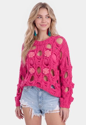 Donna che indossa un maglione a maglia grossa di colore rosa acceso con grandi buchi e dettagli floreali abbinato a dei pantaloncini in denim chiaro strappati.