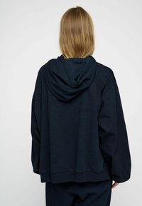 Ciemnoniebieska bluza z kapturem, wykonana z miękkiego materiału, o lu�źnym kroju, z długimi rękawami i obszerną tylną częścią z kapturem. Prosty design bez widocznych elementów metalowych.