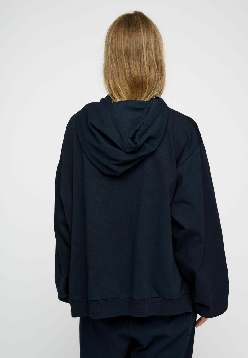 Ciemnoniebieska bluza z kapturem, wykonana z miękkiego materiału, o luźnym kroju, z długimi rękawami i obszerną tylną częścią z kapturem. Prosty design bez widocznych elementów metalowych.