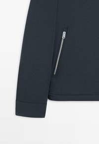 Mørkeblå jakkeærme og nederste sektion med en lodret sølv lynlåslomme på højre side mod en hvid baggrund.