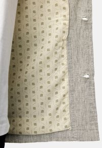 Tissu beige avec de petits motifs géométriques marron à côté d'un tissu gris clair texturé comportant deux boutonnières.