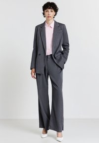 Frau, die einen grauen Nadelstreifenanzug mit weit geschnittenen Hosen und einem rosa Hemd trägt, steht mit einer Hand in der Tasche und weißen Schuhen mit hohen Absätzen.