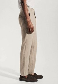 Beige lněné kalhoty s rovnými nohavicemi, vybavené poutky na opasek, postranními kapsami a hladkým povrchem. V kombinaci s tmavě hnědými mokasíny.