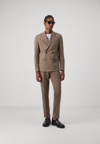 Completo doppiopetto marrone chiaro con una vestibilità su misura, abbinato a una camicia bianca e scarpe nere. Il modello indossa occhiali da sole neri.