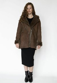 Brun ruskindsjakke med en pelsforet krave, lynlåslukning foran og skrå syninger, kombineret med en sort tætsiddende kjole og ankelstøvler.