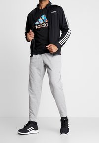 Schwarze Adidas-Jacke mit weißen Streifen, schwarzes Grafik-T-Shirt, graue Jogginghose und schwarze Sneaker mit weißen Akzenten. Lässige Sportbekleidung.