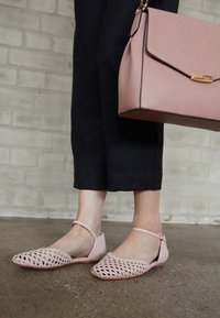 Pieds chaussés de ballerines tissées rose clair avec des brides à la cheville et un pantalon noir, tenant un sac à main assorti rose clair contre un mur en briques grises.