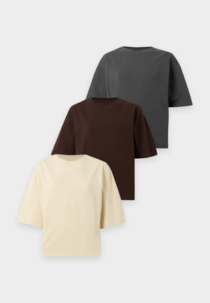 Femme et homme debout côte à côte portant des t-shirts oversize gris foncé avec des bas noirs sur un fond blanc uni.