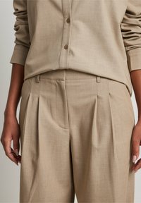 Chemise beige sur mesure et pantalon plissé en tissu texturé, avec une fermeture à boutons et des poches latérales.