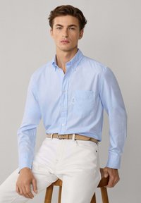 Camisa de botones azul claro con un bolsillo, hecha de algodón, combinada con pantalones blancos y un cinturón marrón. Modelo sentado en un taburete de madera.