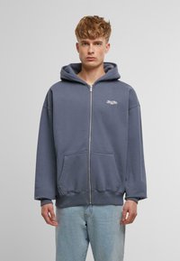 Hooded zip-up sweatshirt in dark gray cotton. Features front pockets, ribbed cuffs, and a small white logo on the chest. Soft texture.