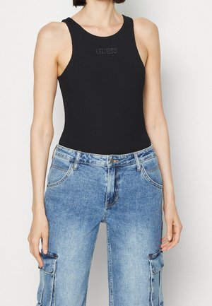 Mujer con una camiseta sin mangas negra con el texto "GUESS" y unos vaqueros cargo de tiro alto en azul claro, delante de un fondo blanco.