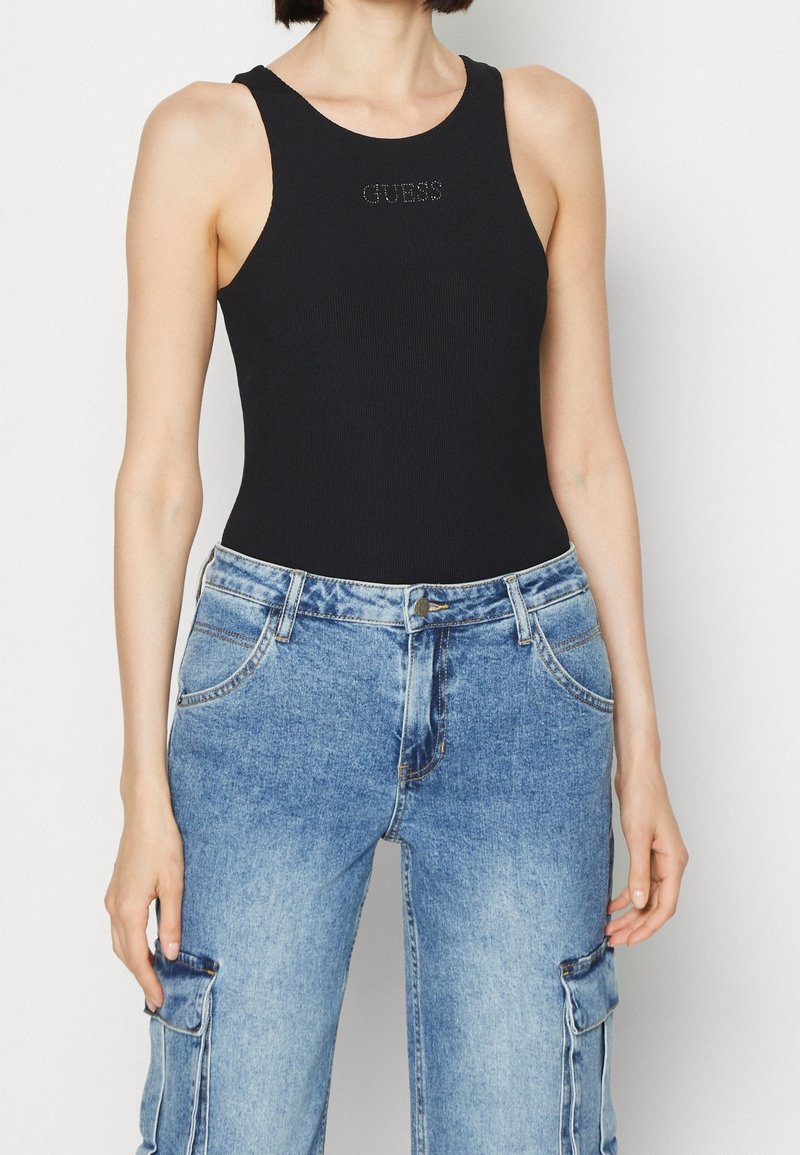 Femme portant un débardeur noir sans manches avec le texte « GUESS » et un jean cargo taille haute bleu clair sur fond blanc.