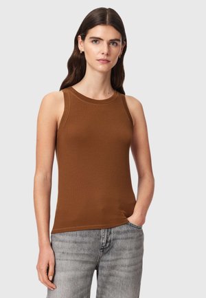 Frau mit langen dunklen Haaren trägt ein ärmelloses braunes Top und hellgraue Jeans, steht mit einer Hand in der Tasche vor einfachem Hintergrund.