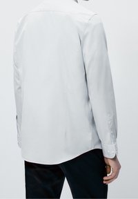 Camicia grigia chiaro a maniche lunghe con polsini a bottoni, texture liscia, orlo posteriore spaccato e colletto classico, vista dal retro.