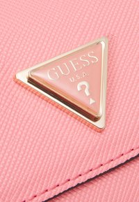 Różowa teksturowana powierzchnia z trójkątnym logo Guess z złotą obwódką, które zawiera napis "GUESS U.S.A." oraz symbol zapytania.