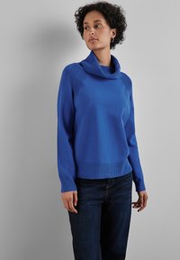 Kobaltblaues Sweatshirt mit einem Schalkragen, strukturierten Schulterakzenten und geripptem Saum. Getragen mit dunklen Jeans.